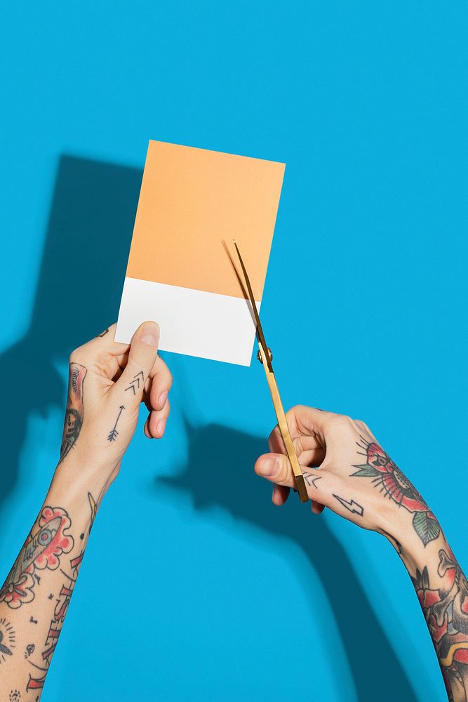 Tattooed hands cutting a yellow paper with a blue wall