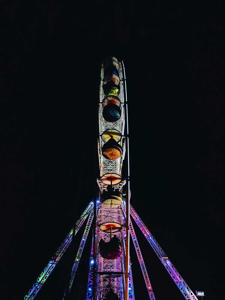 A Ferris wheel at a fairground lit up in bright colors at night. Original public domain image from Wikimedia Commons