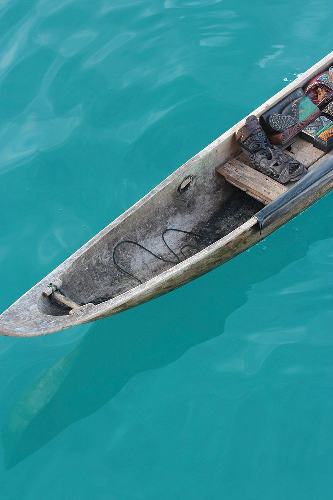 A wooden canoe with tribal masks inside on the azure surface of water. Original public domain image from Wikimedia Commons