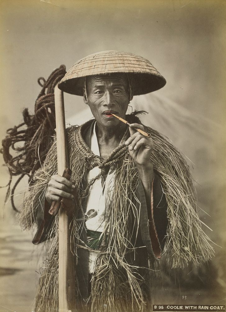 Portrait of a man with wrinkled, leathery skin, wearing a straw hat and a plant fiber cape-like garment; man holds a smoking…