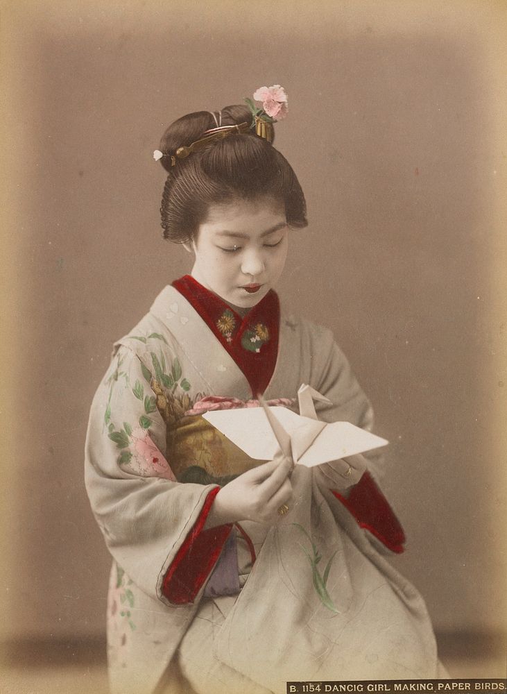 young woman wearing grey kimono with pink and yellow flowers, holding an origami bird made of white paper. Original from the…