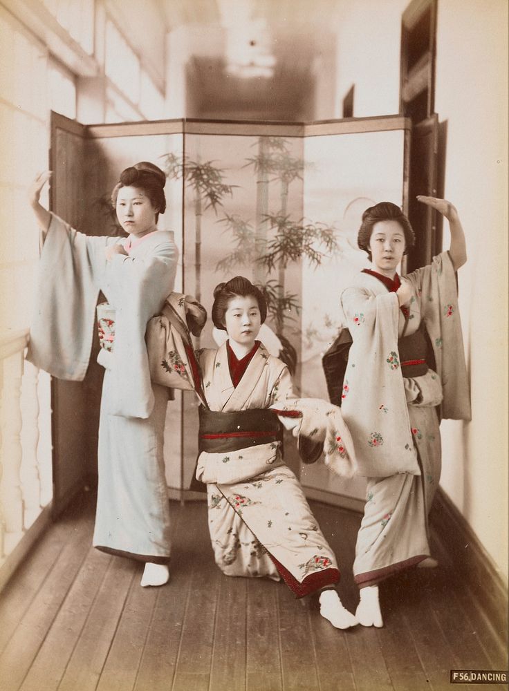 three young women wearing traditional Japanese costumes, in dancing poses, in a narrow hallway in front of a folding screen;…