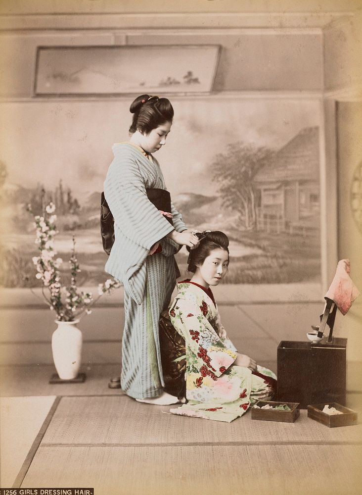 two young women wearing traditional Japanese costumes; seated girl wearing a multicolored flowered kimono with a mirror on a…