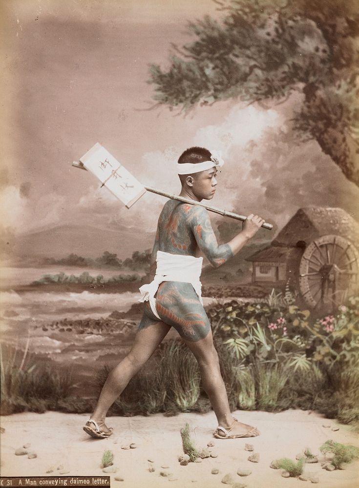 Striding young man wearing white loincloth, white headband and plant fiber sandals, carrying a flat packet attached to a…
