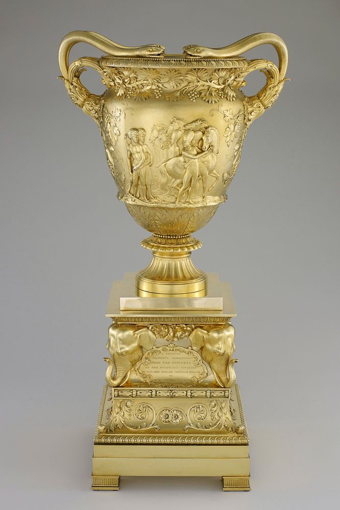 double-handled vase on fluted base; handles are each two snakes with heads resting on rim of vase; relief designs of…