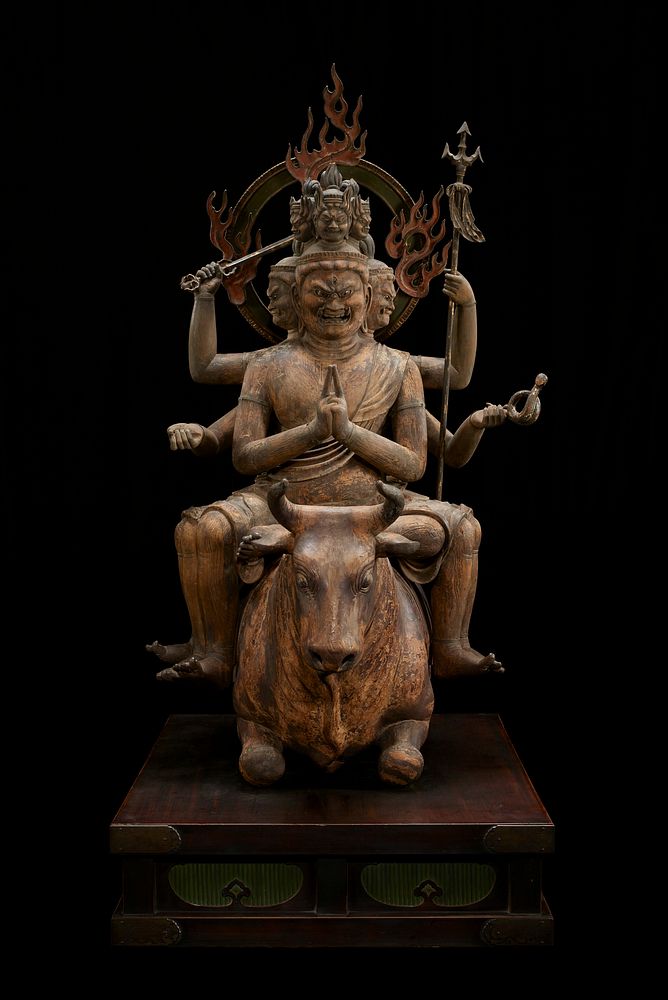 Figure with six heads, arms, and legs astride crouched bull on wood base; metal bands around wrists, some biceps, and most…