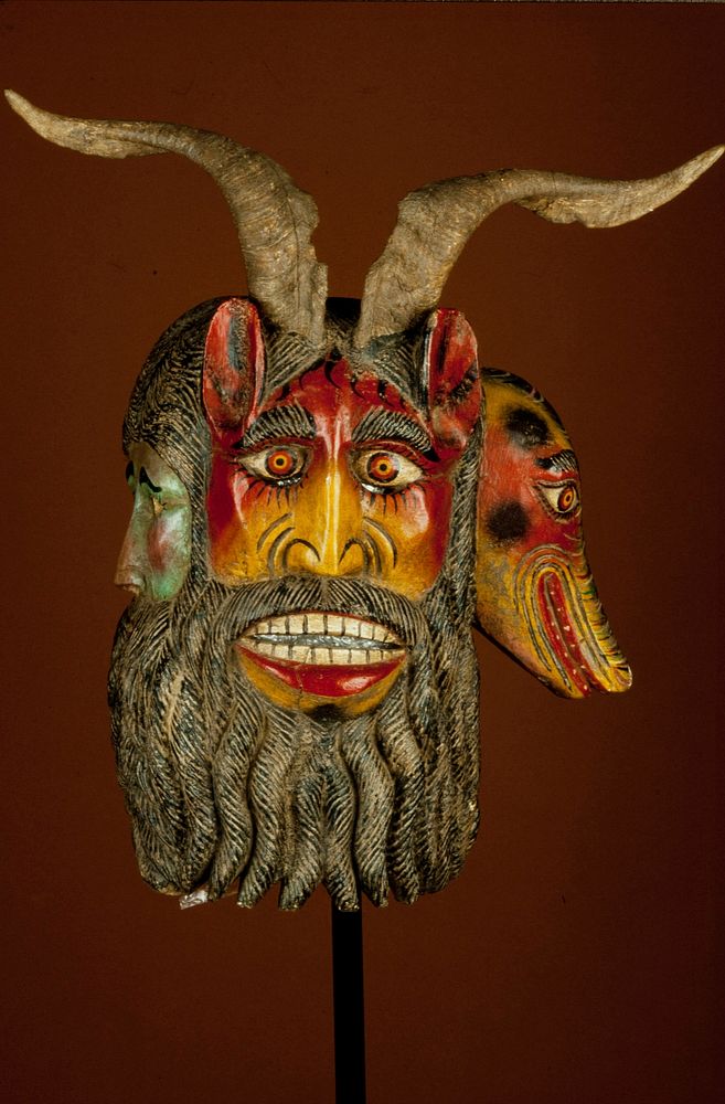 mask, wood, attached goat horns, painted orange, green, brown,red, yellow, black Triple-face mask with human face to left…