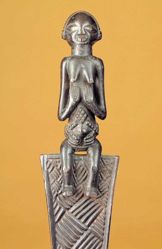 Female figure seated on top of a fan-shaped element decorated with basketweave carvings; small flared area with carved ribs…