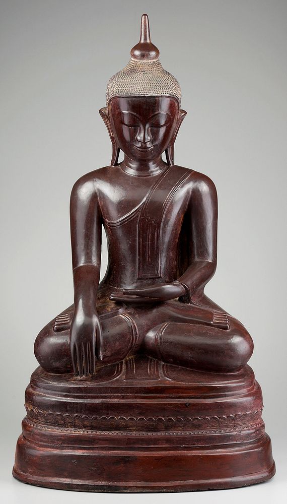 seated cross-legged Buddha with PR hand pointing downward and PL hand palm up on lap; head bent slightly downward, eyes…