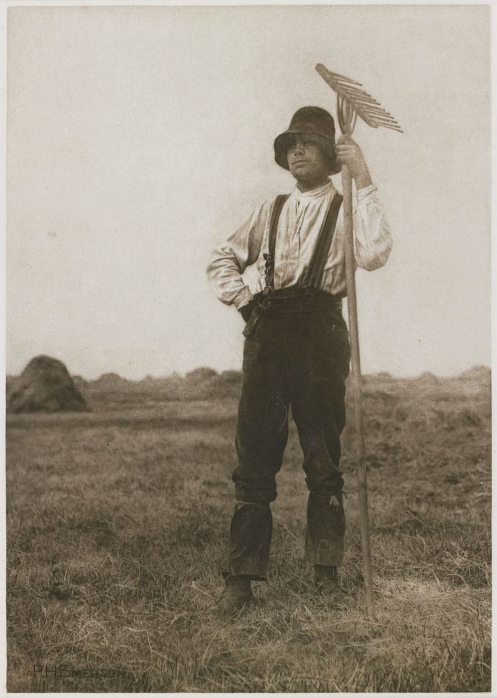 man standing in a field with haystacks in background; man wears a hat, long-sleeved shirt, pants with suspenders and boots…