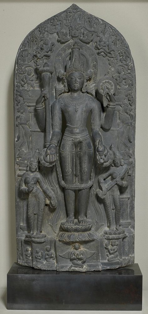 Vishnu on a lotus pedestal flanked by his principal consorts; holds a mace, sun dish (cakra), conch and lotus blossom in his…