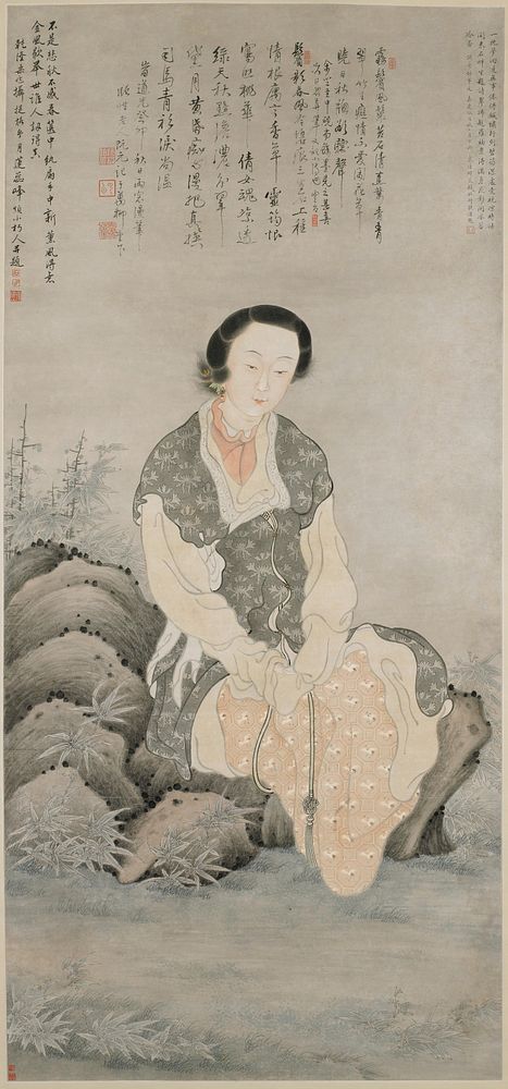 Woman seated on rocks, wearing long vest-like garment in grey with stylized floral designs and a skirt with design of cranes…