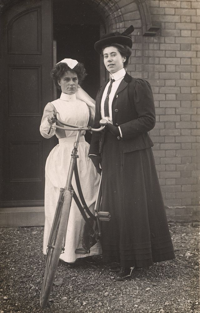 Queen's Nurse. Postcard featuring a black and white photograph of two women standing next to a bicycle. The woman on the…