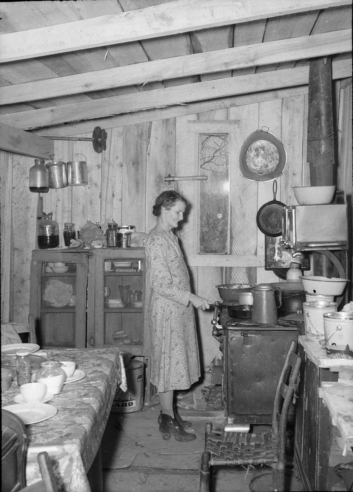 Mrs. Spiller at stove in their one-room shack. Note narrow window. Also Mrs. Spiller wearing overshoes because of dampness…