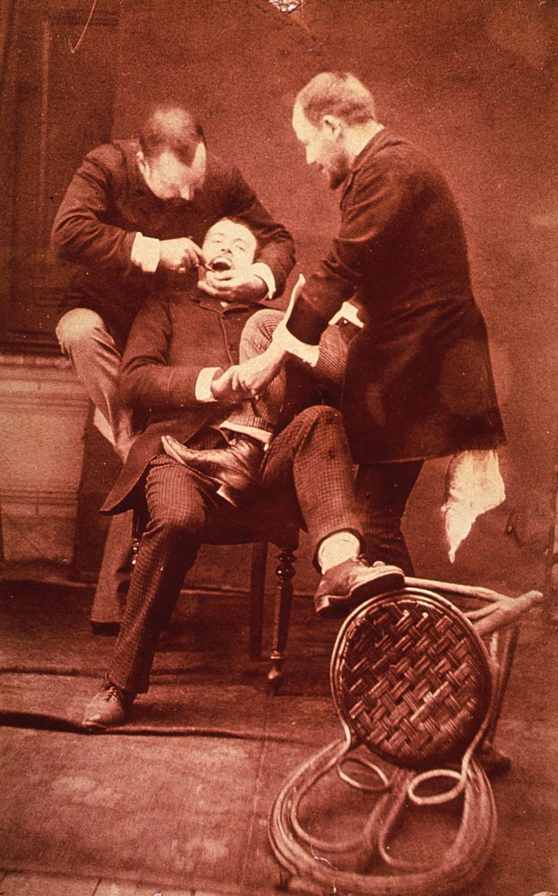 Tooth extraction. A patient is sitting in a chair; a dental assistant has his foot on the patient's lap; behind them the…