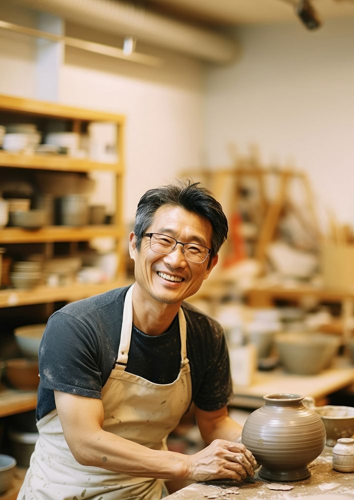 Photo of a thin middle age asian man wearingglasses, casual clothes and apron making pottery. 