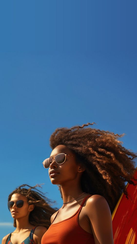 Photo of a black woman carrying surfboard with her friend on the beach, on a blue sky, sunny day.  