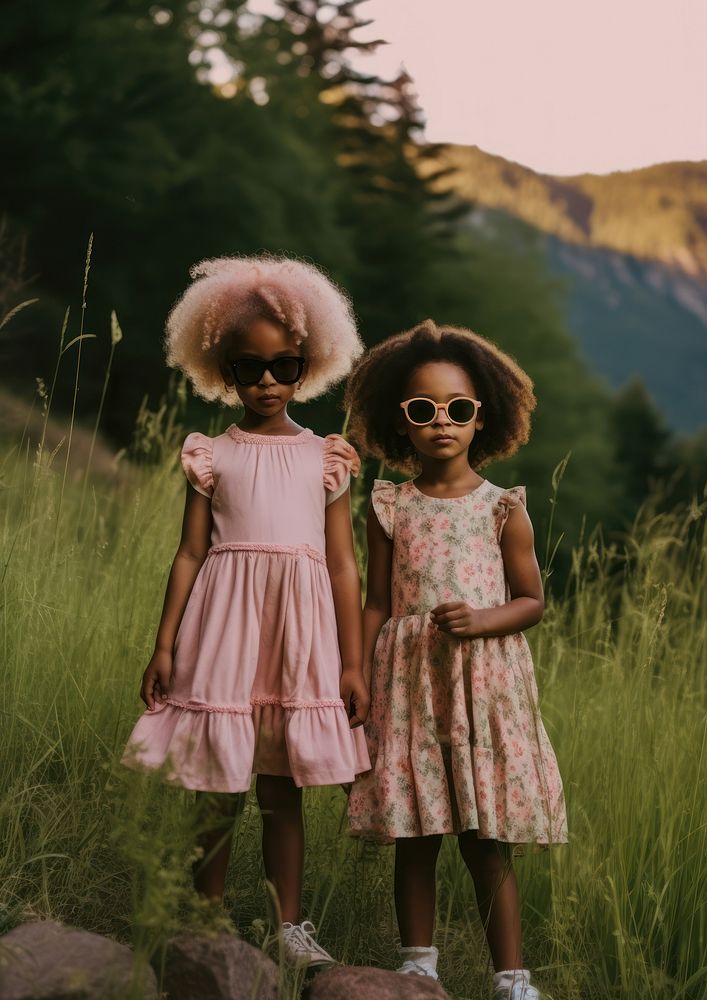 A two black little girl wearing pink dress and cute flowr shape sun-glasses standing next to each other on the green yard in…