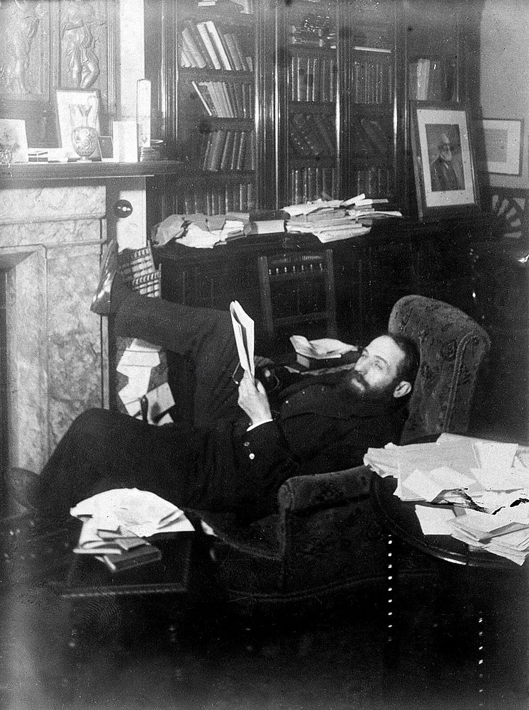 Arthur Schuster, lying on an armchair by a fireplace, with a photograph of Lord Kelvin behind him. Photograph, c. 1901.