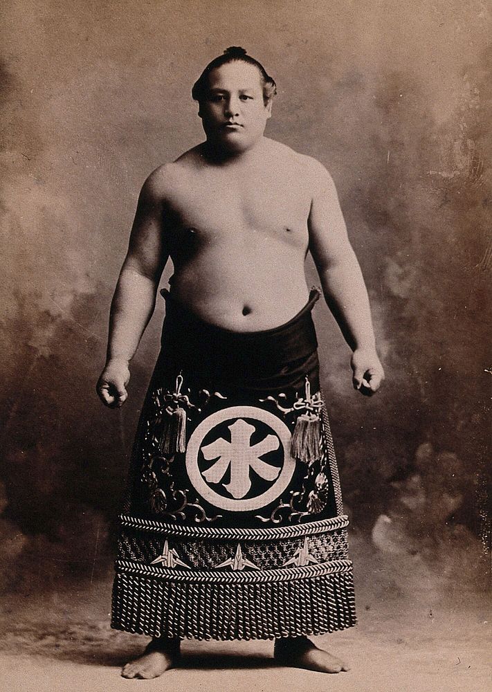 A young sumo wrestler posing in a photographic studio, wearing a richly decorated apron, in front of a painted backdrop.