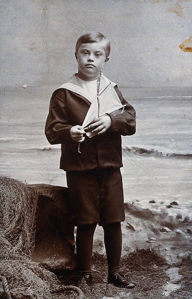 A boy with Down's syndrome, dressed in a sailor suit, standing against a nautical background, holding a small yacht.…