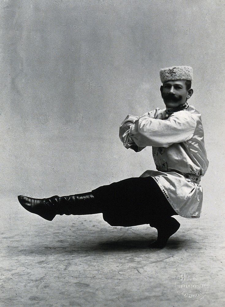 A man, in a satin tunic and fur hat, dancing a "Cossack" measure in a studio setting.
