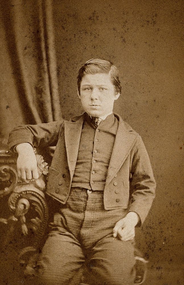 A seated young man with clenched fists, leaning against an ornate table. Photograph by A.E. Scales.