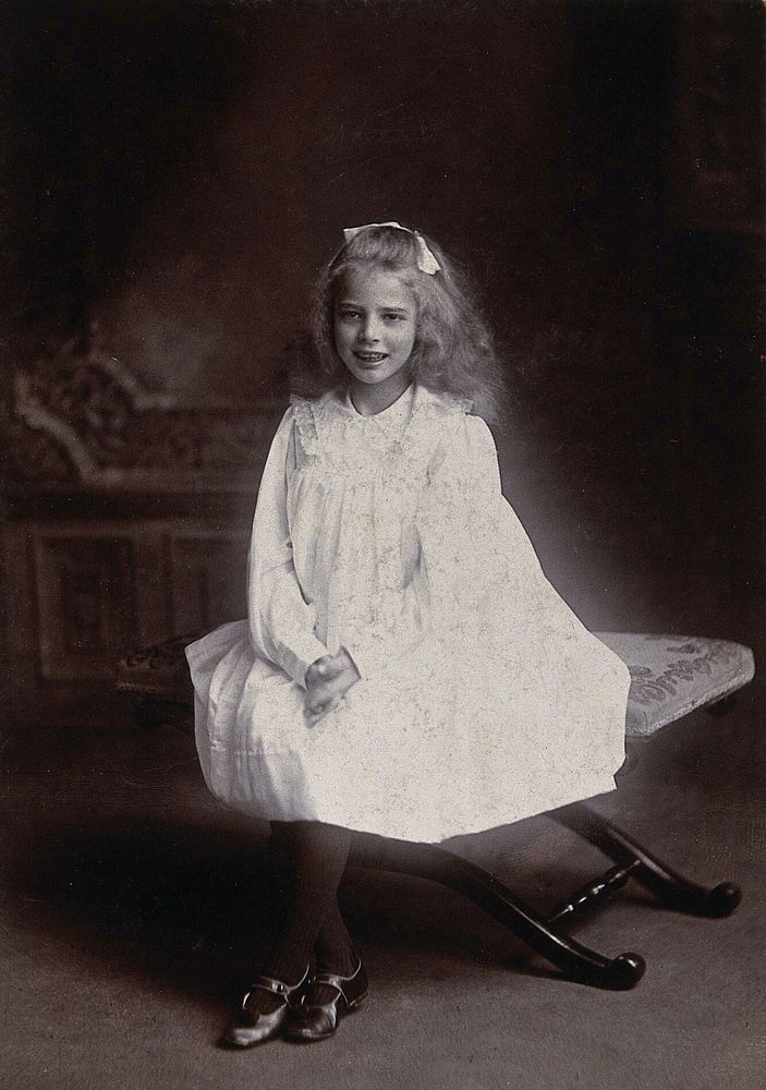 A girl wearing a white dress, sitting on an embroidered stool. Photograph by Lock & Whitfield.