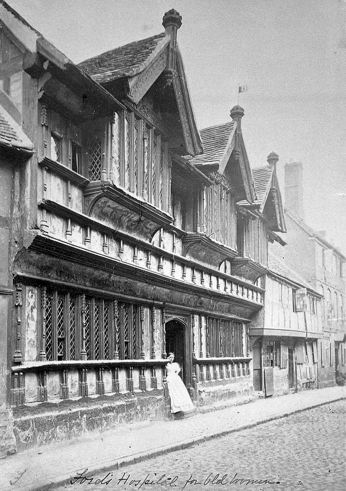 Ford's Hospital, Coventry: exterior from the street, with a woman standing in the doorway. Photograph.