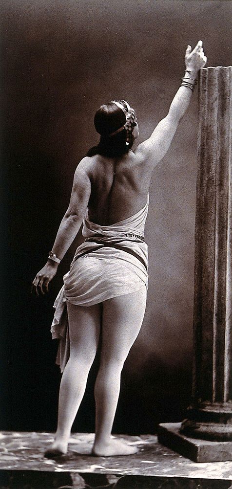 A young woman posing semi-naked, back view, standing on a podium beside a truncated classical column, in a photographic…