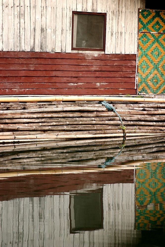 A stacked bundle of bamboo rests below a wooden slatted wall with a small window. The water below reflects it all.