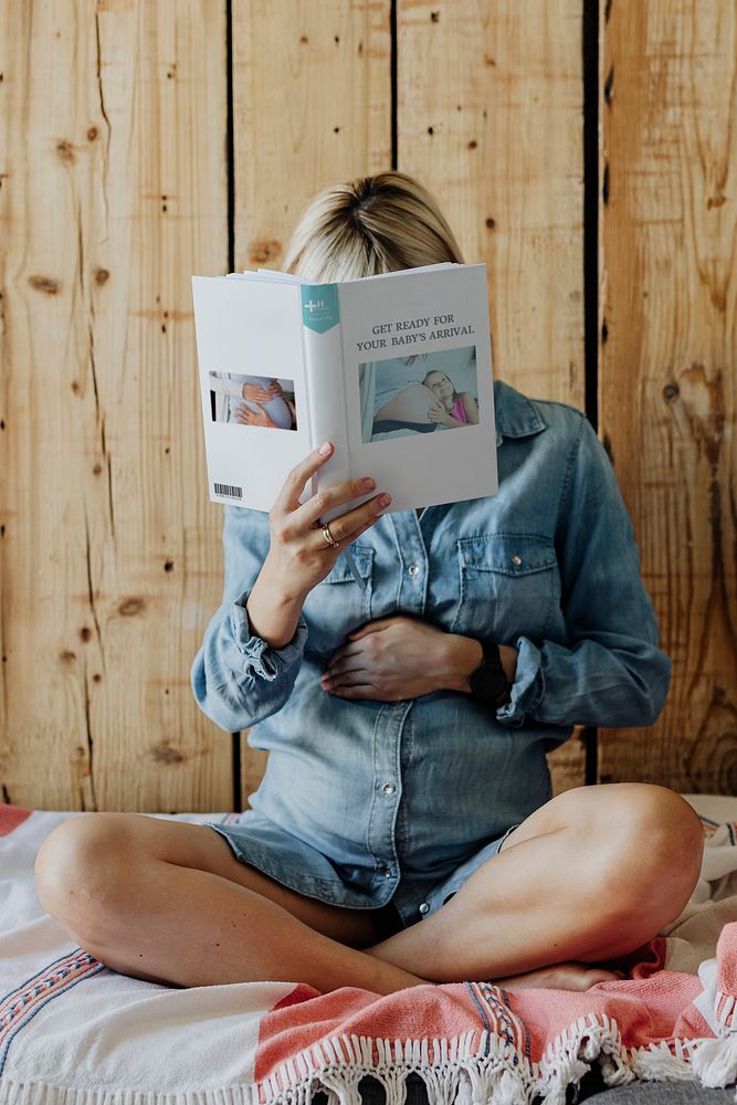 Pregnant woman in a denim dress reading a book on a couch
