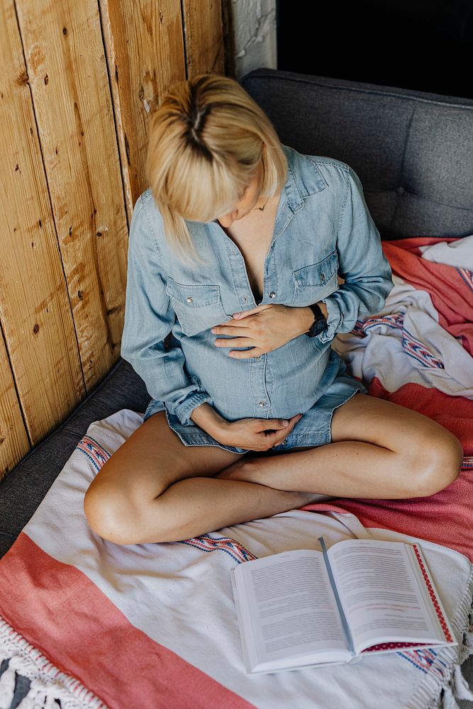 Pregnant woman in a denim dress reading a book on a couch