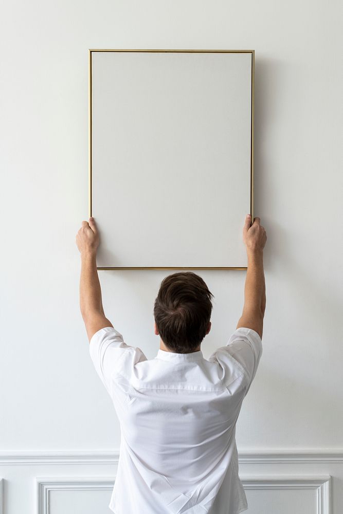 Blank frame being hung by a young man on a white minimal wall