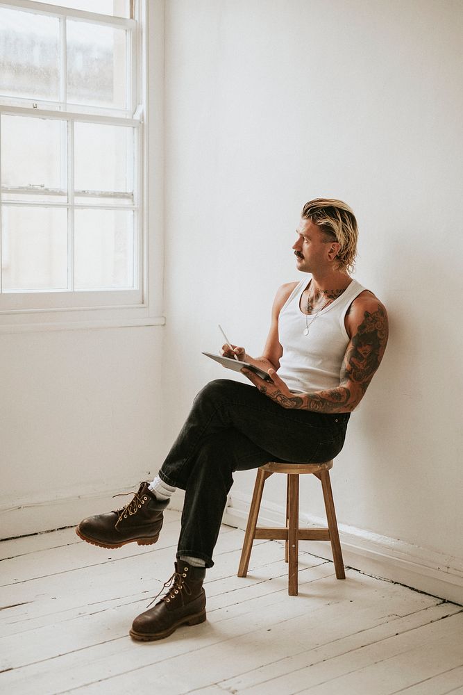 Alternative tattooed man in white tank top using digital tablet