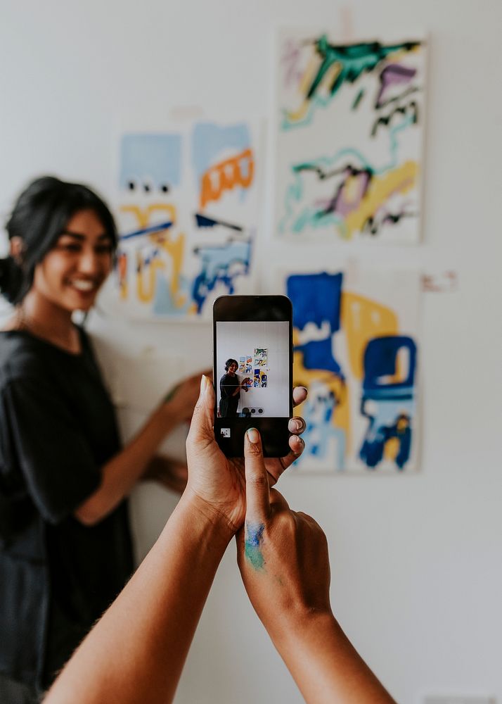 Friend taking a photo with phone camera of her artist friend by a mood board on a white wall