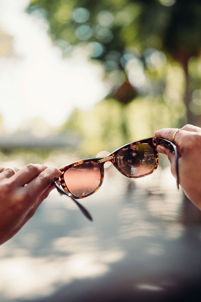 Hands hold up sunglasses with the view of a park blurred in the background.