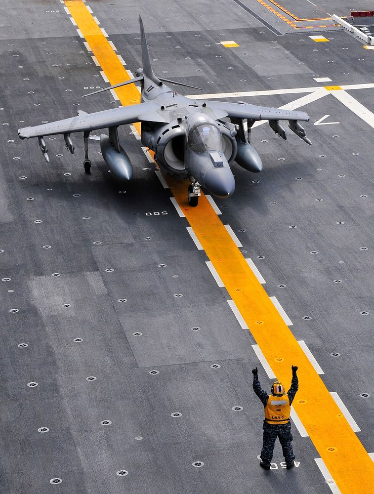 A U.S. Navy aviation boatswain's mate handling taxis an AV-8B Harrier II aircraft on the flight deck of the amphibious…