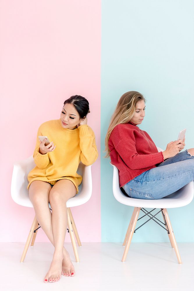 Caucasian and asian women sitting using mobile phones together with pink and mint green background