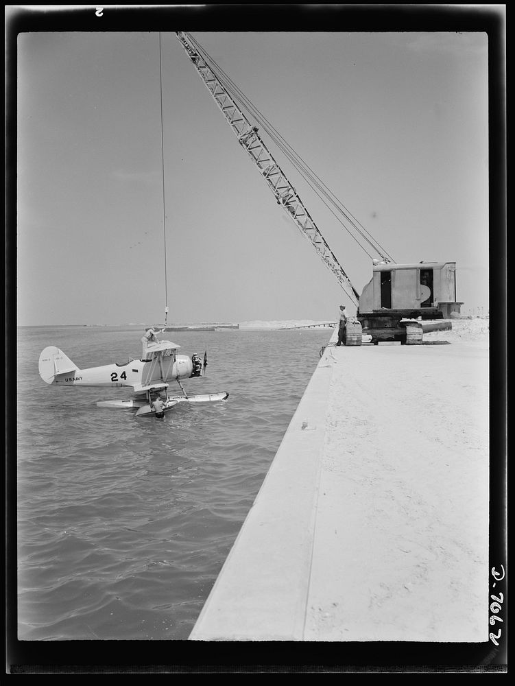 Naval air base, Corpus Christi, Texas. Hooked to a derrick, a Navy N3N plane is made ready for hoisting out to shore onto a…