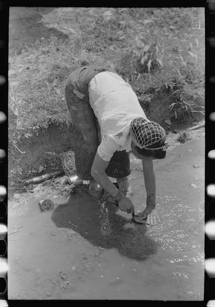 Washing off the plastering trowel in the irrigation ditch, Chamisal, New Mexico by Russell Lee