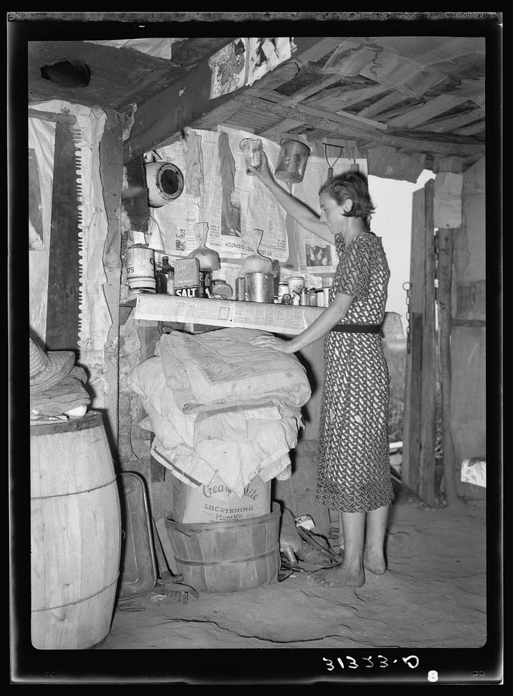 Woman living in shack at Tin Town, Caruthersville, Missouri, in front of shelf used as kitchen cabinet. Note ceiling and…