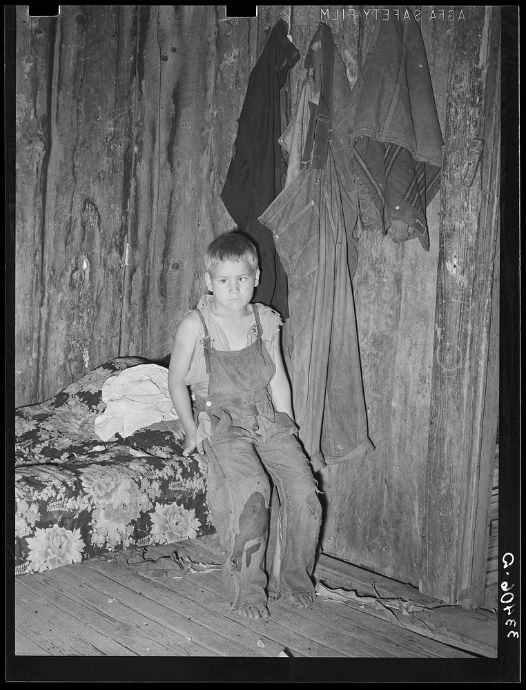 Son of agricultural day laborer in corner of room. The garments hanging on the wall were all the clothes this family of…
