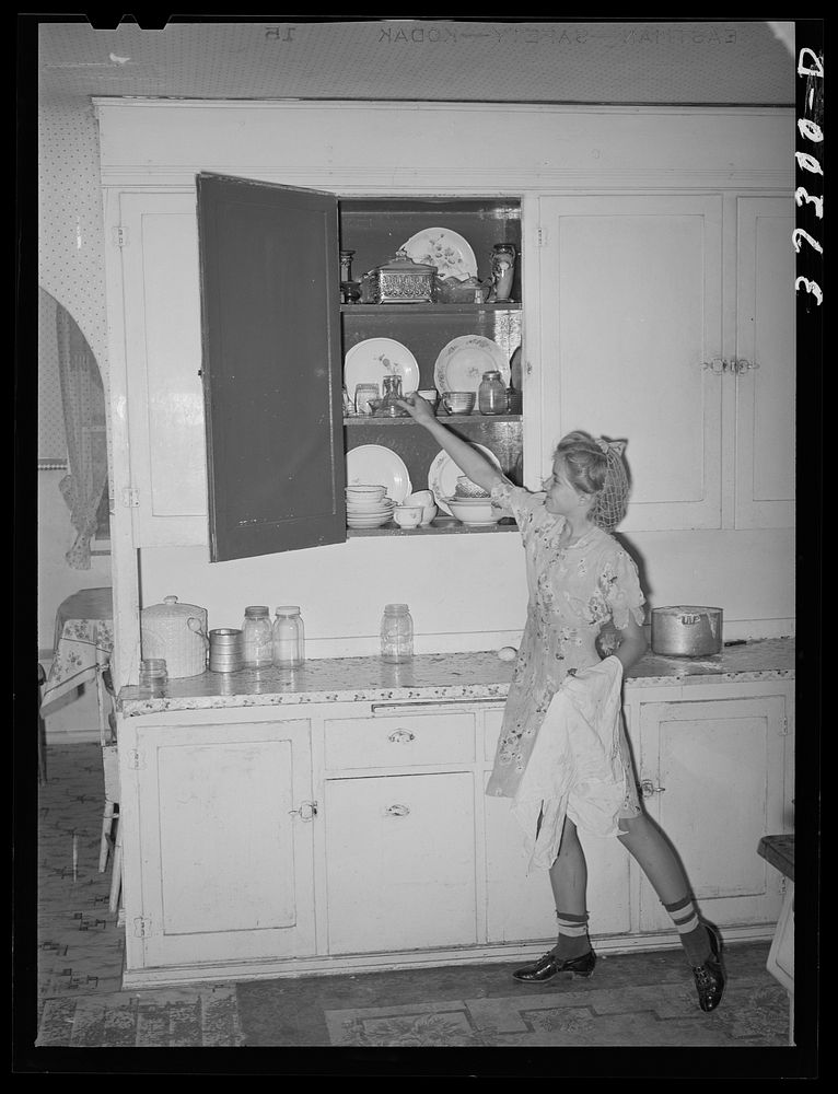 [Untitled photo, possibly related to: Daughter of Morman [i.e. Mormon] farmer putting away dishes in kitchen cabinet. Box…