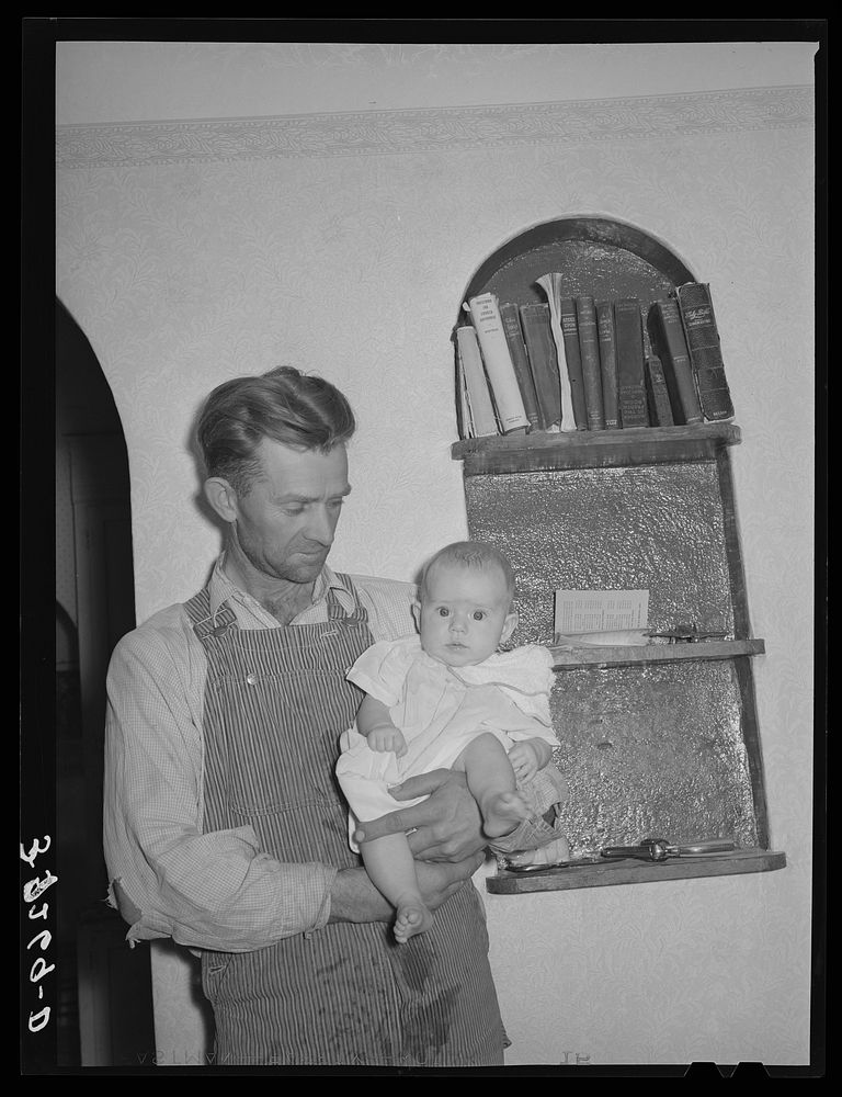 Mormon farmer with his baby in front of a bookshelf in his home. Snowville, Utah by Russell Lee