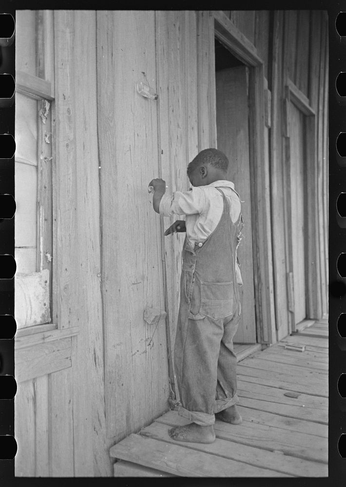 Child of former sharecropper, playing primitive musical instrument made by stretching wire from one nail to another creating…
