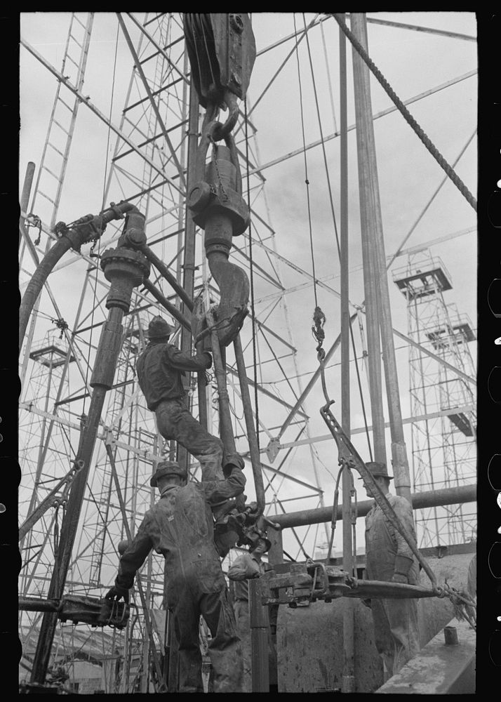 Activity at oil well; man on lengths of traveling block will be pulled up to the crown block at top of derrick, Kilgore…