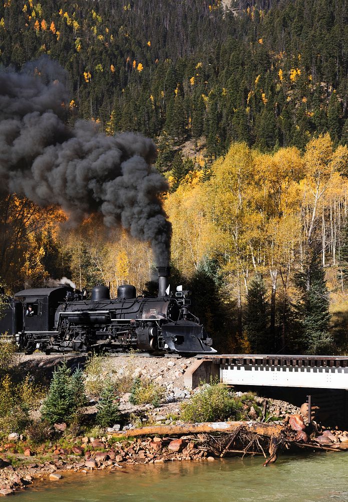 A Durango & Silverton Narrow-Guage Scenic Railroad train, pulled by a vintage steam locomotive, crosses an alpine bridge in…