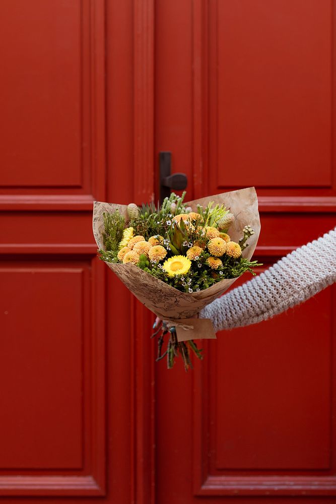 Hand holding a bouquet of assorted yellow flowers in front of a red door