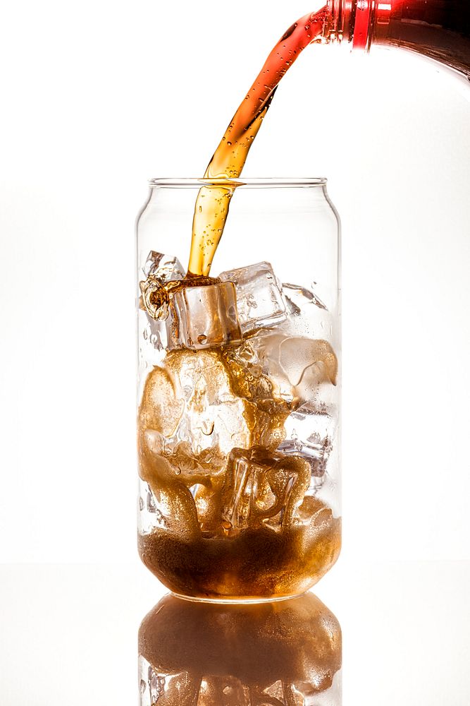 Cold carbonated drink being poured over ice cubes in a can shaped glass 